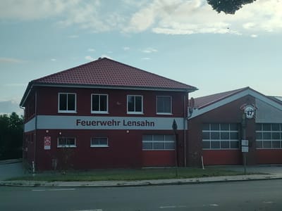 The image shows the fire station in Lensahn, Germany. The building is predominantly red with a red tiled roof. A large white band runs across the front of the building, bearing the text "FEUERWEHR LensaHN" in dark red lettering. The fire station has multiple windows and at least two large bay doors, one of which is visible on the right side of the building and appears to be a garage door with a grid pattern. To the right of the main building, there is an extension with a brick facade. Above the bay door, there is a circular emblem with the number "47" and the word "FEUERWEHR" (fire department) written on it. The building is situated next to a road, with a sidewalk and grassy area in front of it. Trees are visible in the background to the left of the building. The sky above is partly cloudy. There are no people visible in the image.