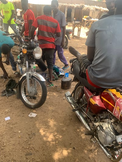 In Gobbiya, Nigeria, a group of men are gathered around two motorcycles in a dusty, outdoor setting. One motorcycle, predominantly silver and red with yellow accents, is in the foreground, viewed from the right side. A person is seated on this motorcycle, with their bare foot resting on the ground and their upper body turned away from the camera. Tools and debris are scattered on the dirt ground around the vehicles.
Behind this motorcycle, another motorcycle, primarily silver with a red seat, is partially visible. Several men are actively engaged with the motorcycles. One man in a bright yellow t-shirt is standing to the left of the silver and red motorcycle, looking down. Another man in a red and black striped t-shirt is standing next to the silver and red motorcycle, appearing to be working on it or observing. A man in a gray and purple striped shirt is standing to the right of the red and black striped t-shirt wearer, looking towards the front. In the background, a simple thatched-roof structure and some wooden tables are visible, suggesting a roadside workshop or market stall. The environment is characterized by dry, sandy earth and sparse vegetation. The overall impression is one of activity and repair work being carried out in a rural or semi-urban Nigerian setting.
