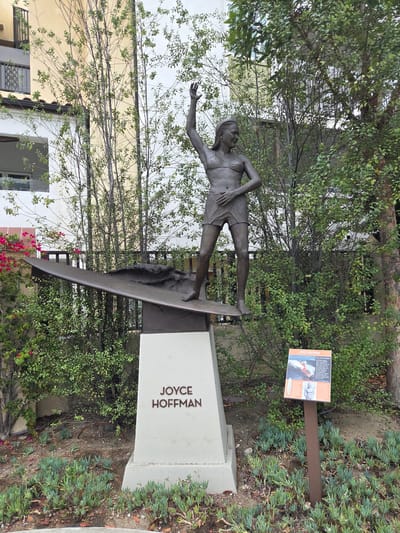 A bronze statue of a female surfer named Joyce Hoffman stands on a surfboard, which is mounted on a white pedestal. The pedestal is inscribed with her name, "JOYCE HOFFMAN". A sign with a picture of Hoffman surfing and biographical information is placed to the right of the pedestal. The statue is located in Dana Point, California, United States of America. The background features a building with white and yellow facade, balconies, and windows, surrounded by lush green trees and plants.
