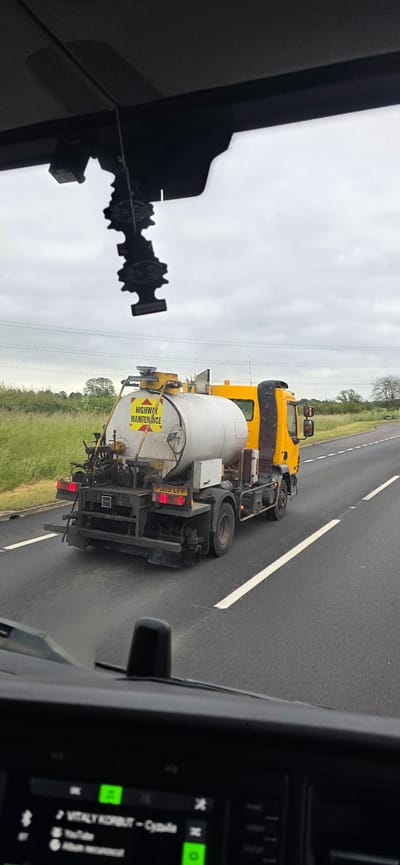 A yellow highway maintenance truck is traveling on a road. The truck has a large white tank on the back. The truck is labeled "HIGHWAY MAINTENANCE". The license plate of the truck is PJ09 LFP. The truck is on a highway with grass and trees on the side. The sky is cloudy. The image appears to be taken from inside another vehicle, as the dashboard and windshield are visible in the foreground. The scene is set in the city of Newark and Sherwood, United Kingdom.
