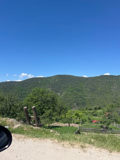 The image shows a landscape in Leshten, Bulgaria, with a clear blue sky and a tree-covered mountain. A road runs alongside the mountain. A wooden structure with two horizontal logs stands next to the road. Some houses with red roofs are visible on the mountainside. The edge of a car mirror appears in the bottom right corner.