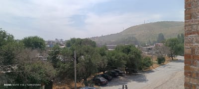 The image is an outdoor, slightly hazy daytime shot taken from an elevated viewpoint, looking towards a semi-arid landscape with a distant hillside. In the foreground on the right, a brick wall is visible. Below the viewpoint, a dirt road curves from the right into the middle ground, with several parked dark-colored sedans lining its side. To the left of the road are numerous green trees and dry, brush-like vegetation. Beyond the trees, several buildings, some with red roofs, are visible, suggesting a semi-urban or village area. In the far distance, a large, barren, and terraced hillside dominates the background, with a few structures and communication towers near its crest. The sky is pale blue with wispy clouds. The image appears to be taken in the city of Kok Jar, Kyrgyzstan, based on the textual overlay in the bottom left corner.