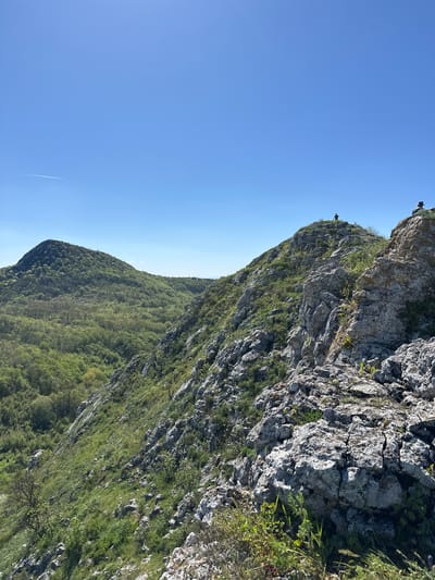 The image depicts a mountain in Bilka, Bulgaria. Two small figures stand atop a rocky cliff at the top of the image. The mountain is covered in dense greenery, transitioning from sparse vegetation on the cliff face to a full forest below. The sky is clear and blue, with a contrail of a plane visible.