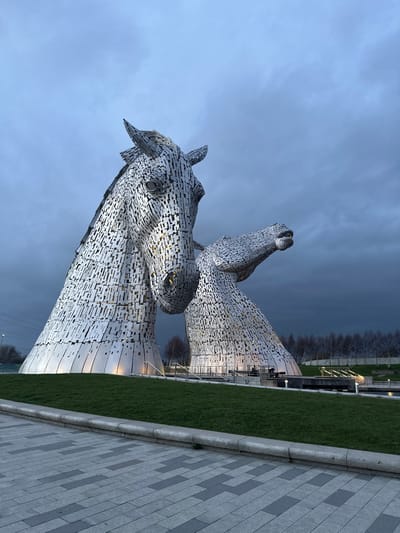 The image displays two colossal equine head sculptures, known as The Kelpies, situated in The Helix park in Falkirk, Scotland, United Kingdom. The sculptures are constructed from numerous interlocking steel plates, creating a textured, metallic surface. The primary sculpture on the left stands upright, its head angled slightly right, featuring visible eyes and nostrils. To its right, the second sculpture's head and neck extend upwards and to the right, conveying a dynamic posture. Both sculptures are illuminated from below, with warm yellow light emanating from their bases and through the gaps in their steel construction. The sky is a dark, overcast blue-grey, suggesting twilight or heavily cloudy conditions. In the immediate foreground, a paved walkway of rectangular light and dark grey blocks borders a strip of vibrant green grass. Behind the sculptures, bare deciduous trees line the distant horizon, and elements of canal infrastructure, including railings and additional lighting, are visible. No human figures are present in the scene.