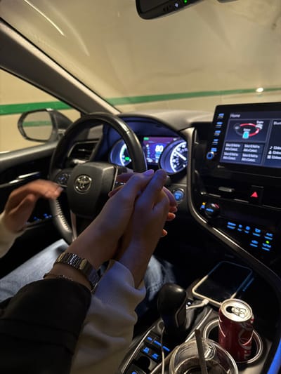 A photograph taken inside of a car shows two people holding hands above the steering wheel. The driver has a silver watch and is wearing a light-colored sweater with a black jacket. The other person has bare hands. The car has a can of Coca-Cola in the cupholder. The make of the car is Toyota. The location appears to be an underground parking garage, although the photo does not allow me to confirm if it is in the city of Grigalati, Georgia.
