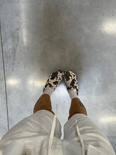 An overhead, medium-level shot captures the lower legs and feet of a person standing on a light grey concrete floor. The person wears white ankle socks and distinctive slip-on shoes with a cow-print pattern, characterized by irregular dark brown or black patches on a cream or white base. Each shoe features a brown strap and buckle detail across the instep. On the right side of the frame, the lower portion of a white garment, possibly shorts or pants with a visible drawstring, is present. The individual's skin tone is medium to dark, and a dark, stylized tattoo is visible on the outer calf of the left leg, just above the ankle. The concrete floor exhibits a fine grain texture with subtle reflections, indicating either an indoor setting or a well-lit covered outdoor area. Minor scuff marks and variations in the concrete's tone are discernible. The image captures a casual moment, emphasizing the person's footwear and lower attire. No text is visible in the image. The location is Su Masu/Elmas, Italy.