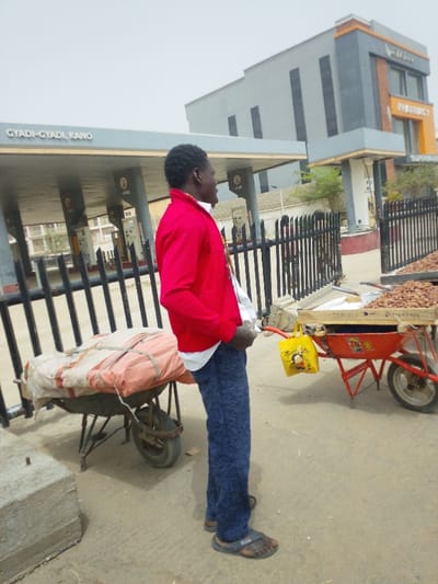 The scene is situated in the city of Daurawa, Nigeria. Visible signage indicates the specific district as Gyadi-Gyadi, Kano.
The image captures an outdoor urban environment under an overcast sky. In the foreground, an adult male stands in profile, facing right. He wears a red long-sleeved jacket over a white shirt, dark blue patterned trousers, and dark flip-flops. A multi-colored lanyard or necklace is visible around his neck. His hands are positioned near his waist.
To the left of the man is a dark-colored wheelbarrow carrying a large, irregularly shaped bundle covered in light-colored fabric and an orange tarp, secured by ropes. To his right, a red wheelbarrow holds a wooden display filled with numerous light-brown, oval-shaped items, consistent with groundnuts or similar produce. A yellow plastic bag with "Thank you" text and a caricature is suspended from the red wheelbarrow's handle.
A black metal fence separates the foreground from the background. Behind the fence, a grey-and-black gas station canopy, marked "GYADI-GYADI, KANO," is visible, with fuel pumps underneath. Further to the right, a modern multi-story building with grey and orange accents features signs for "PHARMACY" and "SUPERMARKET." The ground is paved, suggesting concrete or asphalt. No direct interactions between the man and other individuals or objects are discernible; he appears to be observing something to his right.