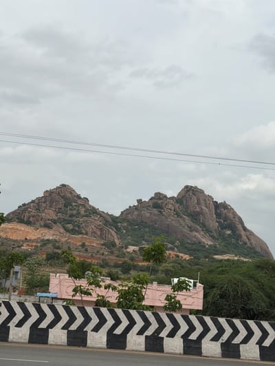 The image shows a cloudy sky over a rugged, rocky mountain range. In the foreground, there is a road with a black and white chevron-patterned barrier. Behind the barrier, there is a pink building with some greenery around it, and further in the background, the mountains rise with patches of green vegetation. There are power lines visible in the sky. The image appears to be taken in Addakal, India, given the context.