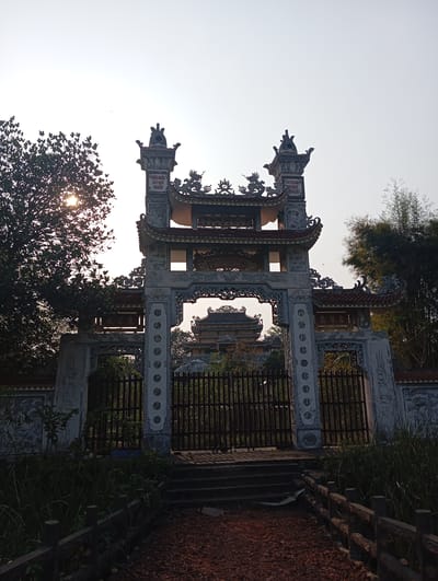 A low-angle, medium shot captures the entrance of a temple in Lumbini Sanskritik, Nepal. An ornate gate stands in the foreground, built of light gray stone, with intricate carvings and inscriptions. It has a red-tiled roof and two vertical inscriptions on either side of the top. A tall, dark wooden gate obstructs access.
Behind the gate, a glimpse of a temple can be seen. To the left of the gate, there is a tree with dense foliage, with sunlight streaming through its branches. To the right, bamboo trees stand tall.
A red gravel path leads up to the gate. Low, wooden fences line both sides of the path. The ground is covered with grass and other vegetation. The sky is a pale gray-white, indicating either early morning or late afternoon.