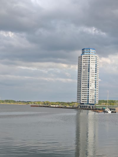 A tall, modern building with white and blue accents stands prominently against a cloudy sky. The building appears to be located on or near a pier or waterway, as evidenced by the surrounding water and docks. A single sailboat is docked nearby, further suggesting a waterfront location. The land in the background is lined with green trees. The reflection of the building shimmers in the water, creating a mirrored effect. The location appears to be near Schleswig, Germany, based on the architectural style and environmental features.