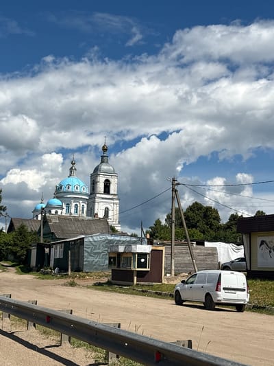 This is a vertical, slightly low-angle photograph of a street scene in a village, likely in Russia, given the provided location context. The primary subjects are a white Lada van parked on a dirt road and a traditional Russian Orthodox church with blue domes. The van is facing away from the camera, and the license plate is partially visible, showing "EOOME 99".
The scene is set on a sunny day with scattered, dramatic white clouds against a bright blue sky. The church, a significant element, features a tall white bell tower with arched windows and a grey dome, topped with a cross. Adjacent to it are smaller buildings, one with distinctive blue domes also topped with crosses. The architecture suggests a historical or religious site.
The road is unpaved and dusty, with a metal guardrail running along the right side of the frame. Several wooden buildings, some appearing weathered and rustic, line the street. The presence of telephone poles and wires indicates infrastructure in the area.
The overall impression is one of a quiet, rural setting on a clear day. There are no people visible in the photograph, and the focus is on the architecture and the natural elements. The lighting suggests it is daytime, likely late morning or afternoon. The image conveys a sense of peacefulness and perhaps a touch of historical charm due to the presence of the church.