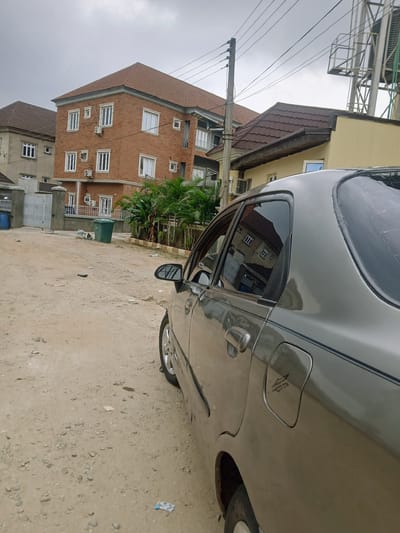The image shows a portion of a gray car parked on a dirt road in Jiru, Nigeria. Adjacent to the road is a building complex with brick and yellow-painted sections, featuring windows, balconies, and a red-tiled roof. Palm trees and a gate are visible in front of the building. Overhead, there are utility poles and wires. The sky appears overcast.