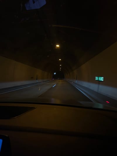 The image depicts the interior of a vehicle driving through a tunnel. The perspective is from the driver's seat, with the dashboard visible in the foreground. The tunnel walls are illuminated by overhead lights, casting a warm, yellowish glow. The road ahead is dark, with lane markings faintly visible. There are some lights on the tunnel walls and on the road itself, likely indicating traffic or safety signals. The overall impression is one of enclosed darkness with artificial lighting. The tunnel is likely part of a road network. The prompt does not provide sufficient information to determine the specific location in the city of Motnik, Slovenia.