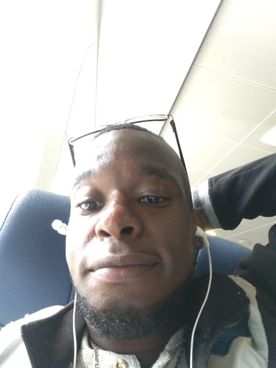 A young man with a dark complexion and short black hair sits on a seat in a vehicle. He is wearing a black jacket over a white shirt. His black framed glasses are resting on top of his head. He has a small goatee and appears to be wearing a necklace. The image is taken from a slightly low angle looking upwards at the man. The location is inside a vehicle, likely a ferry or a bus, based on the overhead lighting and the design of the seat.