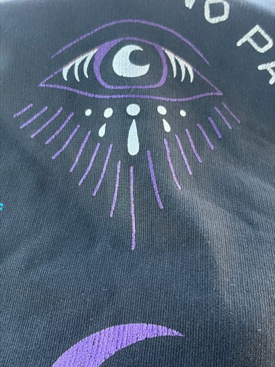 The image shows a close-up of a dark-colored fabric, likely a t-shirt, with a graphic design. The design features a stylized eye with purple and white accents, surrounded by purple lines resembling rays or tears. White dots and a teardrop shape are also present below the eye. Partially visible text in white letters, with a purple outline, is present at the top right of the image. The fabric has a textured knit pattern. The location in the city of Zarech'e, Russia, is not discernible from the image.