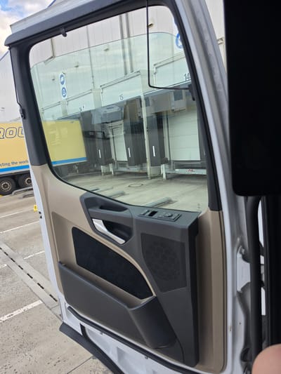 The image captures the interior of a vehicle, likely a truck or van, with the driver's side door open. The perspective is from inside the cabin, looking out through the open door and the side window.

The interior of the door is visible, featuring a dark grey door panel with a speaker grille, a chrome door handle, and a textured fabric insert. The upper part of the doorframe and window seal are also visible, along with the outer body of the vehicle.

Through the open door and the glass, a reflection of the exterior is seen. This reflection shows a row of large white industrial doors, possibly for a warehouse or loading bay, with numbers "14", "15", "16", and "17" painted above them. There are also some signs with symbols, including one depicting a person with a backpack and another with a car and a prohibited sign. In the foreground of the reflection, a yellow truck with blue lettering is partially visible, with its wheels and part of the trailer showing.

The ground outside appears to be a concrete surface, with some white markings. The lighting suggests it is daytime, and the sky is visible in the upper left corner, showing some clouds.

The scene seems to be an industrial or logistics area, given the presence of the warehouse-like structures and the truck. There is no indication of people or any specific activity taking place beyond what is implied by the setting. The overall impression is that of a paused moment, perhaps before or after loading/unloading operations.

The visible text includes numbers on the building ("14", "15", "16", "17") and lettering on the yellow truck, which appears to include "ROL" and "ecting the worl".