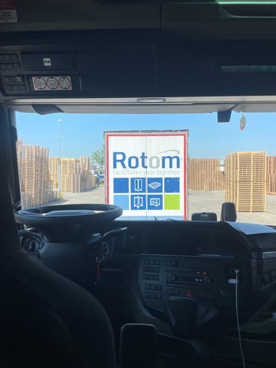 The image is taken from inside the cab of a truck, looking out through the windshield. The scene depicts an outdoor industrial or logistics area, likely a loading dock or storage yard, under a clear, bright sky suggesting daytime and fair weather.

In the foreground, partially visible within the cab, is a steering wheel, the dashboard with various controls and gauges, and a portion of the driver's seat. A blue pen is clipped to the dashboard. On the left side of the cab, there are several control panels and what appear to be informational stickers. One sticker displays a "RIO" logo, and others show circular icons with text below them, including one that reads "4m".

Dominating the view through the windshield is the side of a white truck trailer. Prominently displayed on the trailer is a large logo for "Rotom" with the tagline "facilitates your logistics". The logo features blue lettering and a graphic design with blue and green squares containing icons of logistics-related items like pallets and carts.

Beyond the trailer, stacked high in neat rows, are large quantities of wooden pallets. These stacks of pallets fill much of the background, suggesting a substantial inventory or shipping operation. In the far distance, a few trees and the tops of buildings are visible, along with what appears to be a light pole. A small, circular cartoon-like sticker with a face is attached to the exterior of the truck cab, visible on the right side of the windshield.

The overall impression is a snapshot of a logistics operation, likely during a delivery or pick-up, in Brachterbeek, Netherlands. The bright lighting and clear sky indicate a pleasant day for outdoor work.