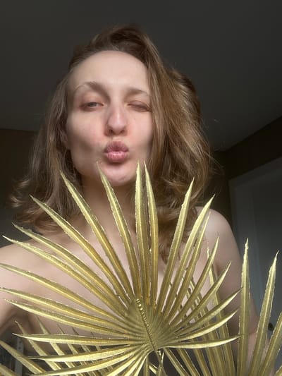 A woman is positioned in front of a large, gold-colored palm leaf, obscuring part of her body. Her face is angled toward the camera with her lips pursed in a kissing gesture. She has light skin, light brown hair, and appears to be indoors in a room with dark, neutral-toned walls. The location details provided (Podshivalovo, Russia) cannot be directly verified from the visual content of the image.