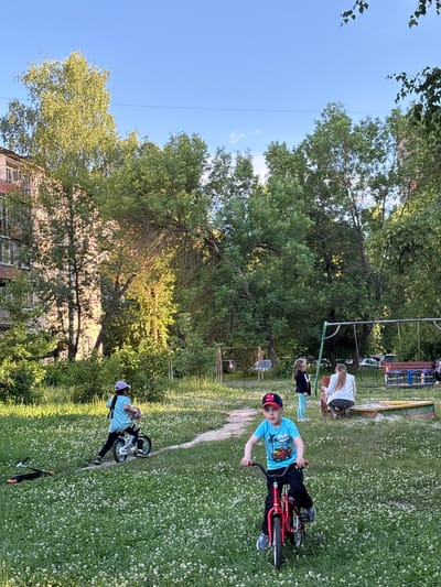 The image depicts a residential courtyard in Sarov, Russia, on a clear, sunny day. In the foreground, a young boy in a blue t-shirt and a red baseball cap rides a red bicycle. To his left, a young girl in a light blue t-shirt, black leggings, and a purple helmet rides a bicycle. A scooter is lying on the ground near the girl. In the background, a playground with swings and a sandbox is visible, with several people present. A woman in a white jacket is sitting near the sandbox, and another young girl in a black jacket and light blue leggings stands nearby. A tall apartment building is situated to the left of the scene, with trees and lush green grass filling the rest of the space. The overall impression is of a typical summer day in a suburban area.