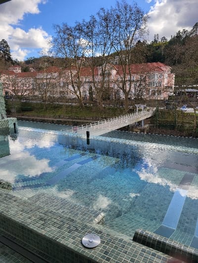 The image displays an outdoor scene in São Pedro do Sul, Portugal, under a clear blue sky with scattered white cumulus clouds. In the foreground, a multi-tiered water feature or pool is visible, featuring greenish-blue mosaic tiles and clear water that reflects the sky. Submerged steps are apparent within this structure. A circular silver fixture is mounted on the rim of the pool. Immediately beyond this water feature, a river or canal flows. Spanning this water body is a white pedestrian bridge with white railings. On the opposite bank, a strip of grass and a row of leafless deciduous trees line the area in front of a large, multi-story building. This building features a light pink facade, white architectural details, multiple windows, balconies, and red-tiled roofs. The structure is set against a backdrop of a densely forested hillside that rises into the background. No individuals or specific actions are depicted.