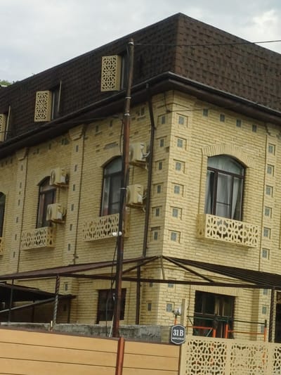 This image captures the exterior of a two-story building constructed with light yellow bricks, located in Kazachiy Brod, Russia. The building features a dark brown asphalt shingle roof with a distinct pyramid-like shape on the upper section. Decorative elements are integrated into the architecture, including ornamental panels above some windows and intricate latticework on small balconies.
The facade is adorned with several air conditioning units mounted on the walls. Windows on the building vary in style, with some being arched and others rectangular. Below the second-story windows, decorative balustrades with a geometric pattern are visible. The building's exterior is also characterized by vertical dark grey rectangular accents and small square windows.
In the foreground, a dark brown support pole stands vertically, with wiring attached to it. Below the second story, a covered outdoor area with a dark brown metal frame and a sloped roof is present. A light wooden fence runs along the bottom of the image. A sign with "31B" and Cyrillic text is affixed to a low wall in front of the building. The sky above is overcast with clouds.