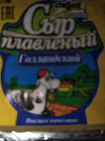 The image shows a close-up of a product label, likely for cheese, with a yellow background. The label features stylized text in Russian and a cartoon illustration of a cow in a rural landscape. The text at the top reads "Сыр" (cheese) and below that "плавленый" (processed). A blue banner displays the text "Галактический" (Galactic). The illustration depicts a cow with a red hat, a house, and green hills. At the bottom of the label, there is faint text that is difficult to read due to the image quality, but it appears to be a slogan or description. There is also a partially visible "EAC" logo on the upper left of the label. The overall image is slightly blurred and of low resolution. There is no indication of any location within the city of Islamabad, Uzbekistan, or any people or interactions.