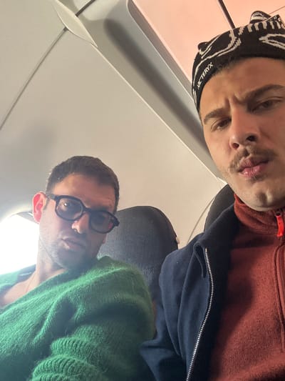 Two men are seated in the cabin of an airplane. The man on the left is wearing a green knitted sweater and black eyeglasses. The man on the right is wearing a black beanie with "ARC'TERYX" written on it, a navy blue jacket, and a rust-colored sweater underneath. The man on the left is looking directly at the camera, while the man on the right is looking off to the right. The coordinates (latitude: 45.613, longitude: 8.716) are in Lonate Pozzolo, Italy, which is near the Milan Malpensa Airport (MXP). The photograph was likely taken inside of an airplane during flight.  There is a small window visible behind the man on the left, showing a bright white light outside. The interior of the aircraft is visible, including the seats, overhead compartments, and a part of the aisle.  The man on the right has a light dusting of facial hair, including a mustache and a goatee.