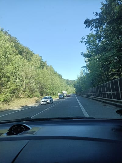 The image shows a view from inside a car driving on a highway lined with trees. There is traffic ahead, including several cars and a truck. The sky is clear and blue. The location is identified as being in the city of Mladá Boleslav, Czechia.