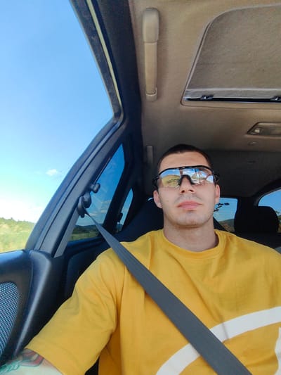 A man wearing a yellow t-shirt and sunglasses is sitting in the driver's seat of a car, with a seatbelt fastened. The car appears to be in motion, as indicated by the blurred scenery visible through the windows and the sky above. The interior of the car includes a sunroof and a grab handle. The man's reflection is visible in his sunglasses, showing a bright sky and trees. The surrounding environment appears to be rural, with rolling hills and vegetation under a clear blue sky. This scene is located in the vicinity of Stefanovo, Bulgaria.
