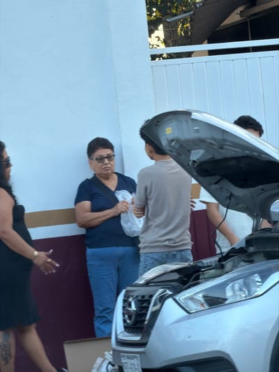 Several people are gathered around a white car with its hood open. An older woman in a blue shirt and jeans is positioned in the center, holding a white cloth. A younger person in a light-colored shirt and jeans stands next to the car. Another person is seen near the front of the car. At the bottom of the frame, a woman in a black dress is lying down. The scene appears to be taking place outdoors, near a white wall.