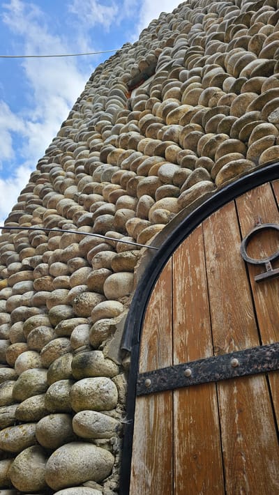 A low-angle, medium shot captures the textured wall of a round building constructed from tightly packed, smooth, gray and tan river stones. The wall extends upwards and to the left, meeting a cloudy, pale blue sky. A thin, off-white wire stretches horizontally across the upper left portion of the frame, disappearing behind the stone wall. Partially visible on the right side of the image is a large, arched wooden door with weathered, vertical planks and dark metal banding. The door features a decorative circular metal emblem with a horizontal bar. The scene appears to be an exterior architectural detail, possibly in Красный Курган, Russia, given the contextual information.