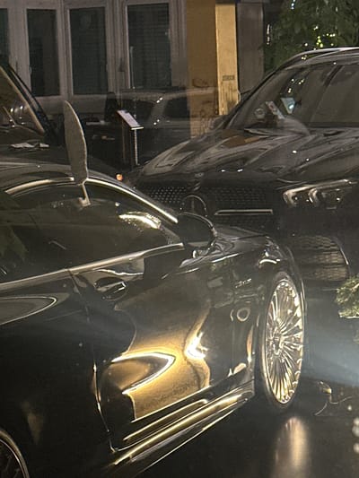 The image captures a black luxury car parked in what appears to be a residential area. Reflected in the car's highly polished surface are the windows of a nearby building, suggesting an urban or suburban environment. The car's front grill and wheel are prominently visible, emphasizing its high-end design. The lighting is low, likely taken at night. The location is likely Wetzikon (ZH), Switzerland, due to the prompt constraints. No people are visible. The focus is solely on the car and its immediate surroundings.