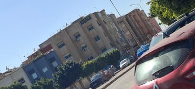 The image depicts a bright, clear daytime scene on an urban street in Berrechid, Morocco, viewed from a slightly tilted, upward-angled perspective. In the immediate foreground, the rear of a dark red hatchback vehicle, identified as a Renault, occupies the lower right portion of the frame, with its rear window and wiper visible.
The street, extending diagonally from the lower left towards the upper right, features several parked vehicles, including a dark SUV, a blue grand taxi, and other passenger cars. On the sidewalk bordering the street, green hedges and small trees are planted, and multiple pedestrians are visible, one wearing a head covering, suggesting routine foot traffic.
Dominating the midground is a multi-story building with a light brown, weathered facade, featuring numerous rectangular windows, some with security bars, and multiple external air conditioning units. A prominent sign with Arabic script is mounted on the building's upper section, reading "مكافحة المخدرات والاتجار غير المشروع بها," which translates to "Fight against drugs and illicit trafficking," indicating a governmental or law enforcement office. To the left of this structure, other multi-story buildings, one with a dark blue facade, are visible. The rooftops of these buildings are extensively covered with various satellite dishes and antennas.
A streetlight pole is visible on the right, and a green pharmacy cross sign is faintly discernible in the upper right background, partially obscured by foliage. The sky overhead is clear and bright blue.
