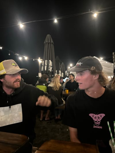 The image depicts a nighttime outdoor social gathering area in Wombarra, Australia. Two adult males are positioned in the foreground, seated at a dark wooden table.

The male on the left wears a beige baseball cap with a yellow mesh back panel and a dark emblem on the front, along with a black hooded sweatshirt featuring a faded white rectangular graphic on the chest. He has dark facial hair, including a beard and mustache, and is looking towards the right side of the frame. His right arm is bent at the elbow, with the hand clenched into a fist, positioned in front of his torso.

The male on the right wears a dark baseball cap and a black t-shirt with a pink graphic on the left chest area, depicting a butterfly with a skull integrated into its body, and the word "ARENDS" printed below. He has light-colored hair styled as a mullet and is looking towards the left, with his mouth slightly ajar. A clear beverage cup containing a green and white striped straw is partially visible on the table to his lower right.

The background, largely out of focus, shows several other individuals seated and standing around additional wooden tables. Multiple large, dark grey outdoor umbrellas are visible, some closed, with partial white text "100...WEEK" discernible on one. A yellow beverage in a glass is present on a table in the mid-ground. The scene is illuminated by numerous overhead string lights with exposed bulbs, and the ground surface appears dark and unpaved.