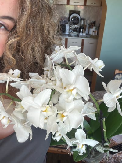The image shows a close-up of a woman's face and a bouquet of white orchids. The woman has curly, highlighted brown hair and is wearing makeup. The orchids are the main subject, with their white petals and yellow centers filling much of the frame. In the background, a kitchen counter with appliances and cabinets is visible. The scene appears to be indoors, possibly a home kitchen, with a pastel blue wall in the background. The overall lighting suggests it is daytime. The location is stated to be Lyubimets, Bulgaria.