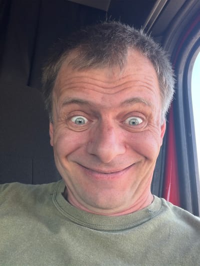 A close-up selfie shows a middle-aged Caucasian man with a wide, somewhat manic grin and his eyes wide open. His hair is messy and graying. He is wearing a green t-shirt. The background suggests he is inside a vehicle, with the dark interior of a cab visible on the left and a window with a red frame on the right. The image was taken outdoors during daylight. The location is the city of North Platte, United States of America.
