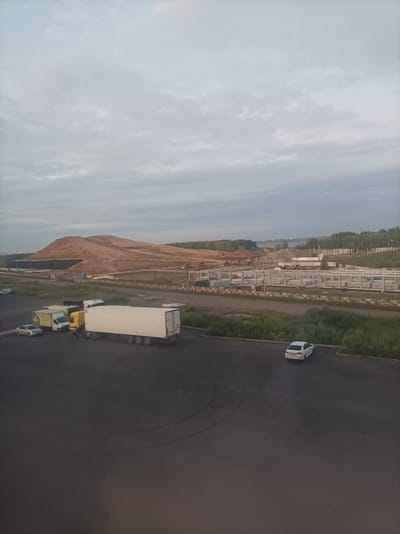 This media file depicts an outdoor scene in Zapadny, Russia, likely during the early morning or late evening due to the overcast sky and soft, diffused light. The sky is filled with a muted palette of grays and whites, suggesting cloudy or dawn/dusk conditions.

The foreground features a dark asphalt surface, possibly a parking lot or industrial area, with tire marks visible. Several vehicles are present. A large, white semi-trailer truck with a yellow cab is parked. Beside it, a smaller yellow truck and a white van are positioned. A silver sedan is parked to the left of these vehicles, and a white sedan is parked further away in the midground. Two figures, appearing to be people, stand near the yellow truck.

The midground and background reveal an ongoing construction project. Numerous concrete pillars and pre-fabricated concrete sections are laid out, indicating the foundation or framework for new structures. Beyond the construction site, a large pile of reddish-brown earth is prominent, possibly excavated material or part of a landscaping effort. Further in the distance, a dense line of green trees borders the landscape, and beyond that, a body of water can be seen reflecting the sky. Construction equipment, including yellow excavators, are visible scattered across the construction area. The overall impression is one of industrial activity and development in a somewhat rural or semi-urban setting. No visible text is discernible in the image.
