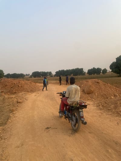 A man sits on a parked motorbike on a dirt road. He wears a light-colored shirt and red pants. Behind him, on the left side of the image, are three people standing in a field. The sky is overcast. Trees line the field's edge. The dirt road takes up most of the foreground of the image.