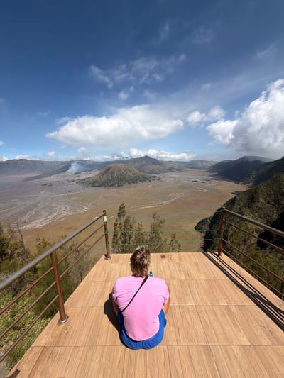 The image depicts a person sitting on a wooden platform, facing a vast volcanic landscape. The person, viewed from behind, is wearing a pink t-shirt and blue shorts. They are seated facing away from the camera, looking out at the valley. The platform is made of light-colored wooden planks and has a metal railing on the sides.
The landscape beyond the platform is a wide, arid valley with a meandering dry riverbed or sand flow. In the distance, there are several volcanic cones, one of which is emitting a plume of smoke. The terrain is characterized by barren, earthy tones with sparse vegetation on the surrounding hills. The sky above is blue with scattered white clouds.
This scene is characteristic of the viewpoint overlooking the Sea of Sand at Mount Bromo, near the village of Cemorolawang, Indonesia. Cemorolawang is a popular tourist destination known for its access to the Bromo Tengger Semeru National Park, offering panoramic views of the volcanic complex.