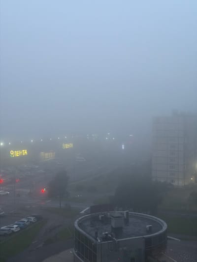 The image depicts a foggy urban landscape, likely in Telmana, Russia. The fog significantly reduces visibility, obscuring distant buildings and details. In the foreground, a circular, modern building with a dark, wet rooftop is visible, featuring visible air conditioning units and other rooftop fixtures. Adjacent to this, a multi-story residential building, appearing to be a Soviet-era panel building, stands to the right.
Further into the scene, through the dense fog, illuminated signs for "ЛЕНТА" (Lenta) are discernible, indicating a commercial area, possibly a shopping center or supermarket. Streetlights and traffic lights are also visible, with one traffic light showing red and green signals. Several cars are parked in a lot and on a street, partially obscured by the fog. The overall atmosphere is dim and overcast, characteristic of a foggy or heavily overcast day. No people are clearly identifiable in the image.