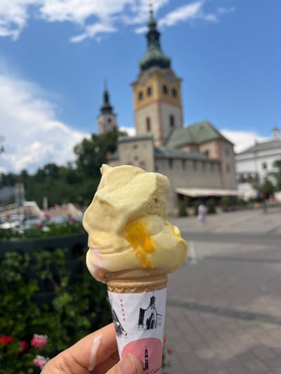 This media file shows a close-up of an ice cream cone held in a person's hand, with a blurred background of Banská Bystrica, Slovakia. The ice cream appears to be light yellow, possibly vanilla or lemon flavored, with some streaks of orange, suggesting a swirl or marbled effect. The cone wrapper has a pink and white design with small circular dots and a silhouette of a building with a steeple.
The background features a cloudy blue sky and architectural elements of buildings in Banská Bystrica, including a prominent tower with a green spire. The buildings appear to be historic, with light-colored facades and arched windows. Green trees and some pink flowers are visible in the lower part of the background, suggesting an outdoor setting with vegetation.
The image is captured from a low angle, looking up towards the sky and the buildings. The lighting indicates it is daytime, likely sunny given the bright sky and shadows. The overall mood conveyed is one of leisure and enjoyment, with the ice cream suggesting a pleasant summer day. There is some melted ice cream dripping onto the person's hand, a common occurrence on a warm day. No specific activity or event is depicted other than enjoying an ice cream.