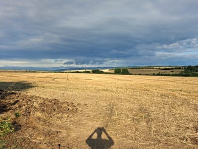 The image depicts a harvested field with dry straw scattered across it. In the foreground, a patch of dark, tilled soil is visible on the left. The horizon is marked by a line of trees and rolling hills under a dramatic sky filled with dark, heavy clouds. A shadow of a person holding a camera is cast on the ground in the middle of the frame. The location is identified as Goran, Bulgaria.