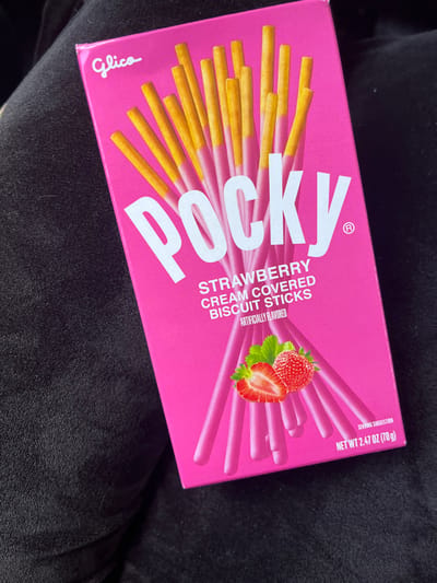 A single box of Glico Pocky Strawberry Cream Covered Biscuit Sticks is depicted, centered and angled on a dark, textured fabric surface. The box is predominantly hot pink with the "Glico" logo in white script at the top left and "POCKY" in large white capital letters across the upper half. Below this, "STRAWBERRY CREAM COVERED BISCUIT STICKS" and "ARTIFICIALLY FLAVORED" are printed in smaller white text. An illustration of two strawberries, one whole and one halved, is located on the lower front. The net weight, "2.47 OZ (70g)," is visible in the bottom right corner. The box features an image of multiple yellow biscuit sticks partially coated in pink cream. The dark background appears to be a soft material, possibly clothing or upholstery, with visible folds and minor lint. No individuals, direct actions, or interactions are visible. The specific location within the city of Edmond, United States, cannot be determined from the visual information presented.