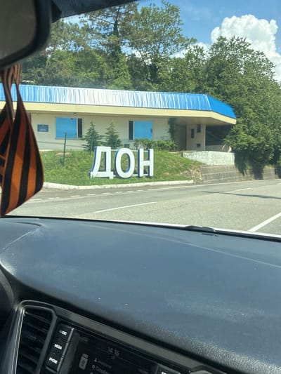 The image shows a view from inside a vehicle, looking through the windshield at a building with a blue roof and the Cyrillic letters "ДОН" in large white block letters on a grassy slope. To the left of the letters, a small blue sign with Cyrillic text is visible on the building. A St. George's ribbon, an orange and black striped ribbon, hangs from the rearview mirror, obscuring part of the view. The building appears to be a roadside structure, possibly an administrative building or a service station, situated behind a low green fence and a retaining wall. Lush green trees and vegetation form a backdrop to the building, under a bright blue sky with white clouds. The dashboard of the vehicle, including parts of the radio and buttons labeled "RADIO," "MEDIA," and "PHONE," are visible in the foreground. The context of the location being Vostok, Russia, is provided as a factual detail.