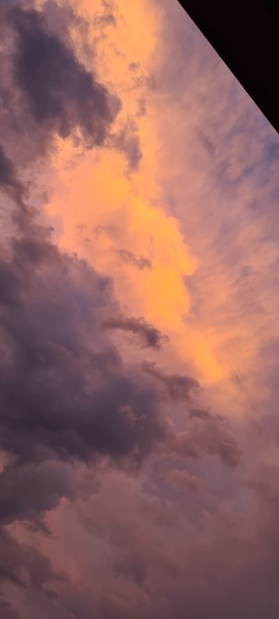 The image depicts a dramatic sky filled with clouds at sunset. The clouds are a mixture of vibrant orange and yellow hues where the sun's rays are catching them, transitioning to darker shades of purple and grey further away. These colors suggest the end of the day and a possibly stormy or overcast weather condition. The clouds are textured and varied, with some appearing wispy and others thick and dense. In the upper left corner of the image, a dark, diagonal shape cuts into the sky, likely representing a building or structure casting a shadow. This object serves as a visual anchor and provides a contrast to the expansive sky. The overall scene evokes a sense of awe and the powerful beauty of nature. There is no visible text in the image.
