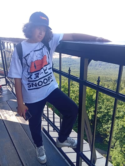A young person with curly hair, wearing a black baseball cap, a white t-shirt with a Snoopy graphic, black pants, and grey and white sneakers, stands on a wooden platform next to a black metal railing. The person is holding a smartphone in their left hand. Their right arm rests on the railing. In the background, a dense forest and a distant landscape are visible under a bright sky. The scene is located on the observation deck of the Sleza Mountain in Sobótka, Poland.