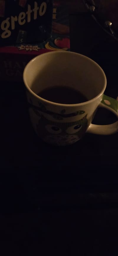 A dimly lit photograph shows a white mug with green and white polka-dot accents on its handle, adorned with green monster illustrations, positioned near the edge of a dark surface. The mug is nearly empty. Next to the mug is a game box, partially visible, titled "Gretto". The background is black, obscuring most details. The location (Gütersloh, Germany) is not discernible from the image.