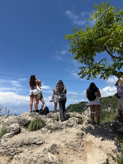 The image shows a group of four people standing on a rocky cliff edge against a backdrop of a blue sky with wispy clouds. In the top left corner, the green foliage of a tree partially obscures the view. The people are all facing away from the camera, appearing to look out at the vista. The location is identified as Pejukutan, Indonesia, suggesting a natural landscape setting. A thin cable runs horizontally across the sky. The rocky cliff is light in color, with sparse vegetation growing on it.