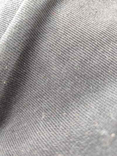 The image is a close-up, abstract view of a dark grey, textured fabric. The fabric has a visible diagonal weave pattern, creating a ribbed effect. There are some small specks of lighter color, possibly lint or dust, scattered across the surface. The lighting creates subtle highlights and shadows that emphasize the texture. The image does not depict any recognizable objects, people, or environmental features that would allow for identification of a specific location within Mizerów, Poland.