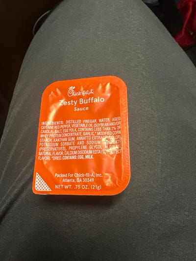 An orange Chick-fil-A Zesty Buffalo Sauce packet rests centrally on a dark grey, textured fabric surface. The packet displays the Chick-fil-A logo, "Zesty Buffalo Sauce," an ingredients list, "Packed For Chick-fil-A, Inc., Atlanta, GA 30349," and a net weight of ".75 OZ. (21g)." A white triangular pattern is visible in the bottom-left corner of the packet. The fabric, likely clothing or upholstery, occupies the majority of the background, with an out-of-focus, darker area visible in the upper-left and a blurred, reddish object in the upper-right. No individuals, specific actions, or interactions are depicted. The precise location in the city of Medford, United States, is not discernible from the visual information provided.